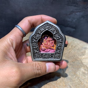May include: A silver-colored, ornate, hinged pendant with a detailed floral design. The pendant contains a small, colorful sculpture of a deity. The sculpture is primarily pink and orange.