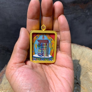 May include: A rectangular pendant with a gold-coloured frame. The pendant features a colourful illustration with red, blue, and yellow hues. The artwork appears to be a religious or spiritual symbol. The pendant is held in a hand.