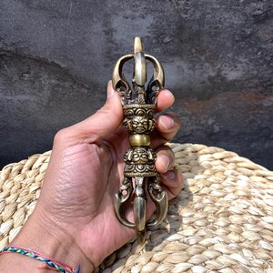 May include: A brass vajra ritual object featuring intricate metalwork, including a central sphere flanked by two detailed faces with expressive features, topped and bottomed by four curved prongs that meet at a single pointed tip on each end.