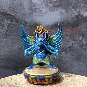 May include: A blue-skinned Garuda statue with large wings, a bird-like face, and a prominent beak. The figure wears a green lower garment, gold jewelry, and stands on a circular base decorated with colorful petals and a gold-toned top surface.