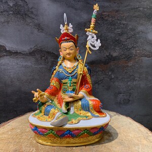 Könnte beinhalten: Eine verzierte Statue einer sitzenden Figur in farbenfrohen Gewändern, einer roten Kopfbedeckung und Goldschmuck. Die Figur hält ein kleines Gefäß und einen Stab mit dekorativen Elementen. Die Statue steht auf einem detaillierten, mehrfarbigen Sockel, vor neutralem Hintergrund.
