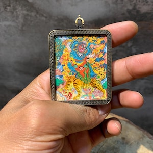 May include: A rectangular pendant with a detailed, colorful illustration of a deity riding a tiger. The artwork features vibrant blues, reds, yellows, and greens, set within a decorative bronze-colored frame. A small gold-colored loop is at the top.