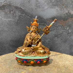 Könnte beinhalten: Goldfarbene Statue einer sitzenden Gottheit mit orangefarbener Krone und aufwendigen Details. Die Statue hält ein kleines Objekt und einen Stab mit dekorativen Elementen. Der Sockel ist mit bunten Mustern verziert, vor strukturiertem Hintergrund.