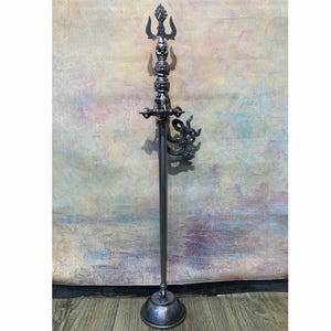 Puede incluir: Un bastón alto de metal oscuro con una punta de tridente de tres ramas, seguido de una pila de tres cráneos humanos esculpidos. Un elemento decorativo de metal en espiral está unido al lado del eje, que descansa sobre una base circular.