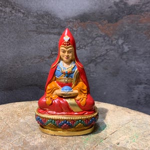 Könnte beinhalten: Eine farbenfrohe Statue einer sitzenden spirituellen Figur. Die Figur trägt einen roten Hut und ein rotes Gewand mit goldenen und blauen Akzenten. Die Statue hält ein blaues Objekt und sitzt auf einem dekorativen Sockel.