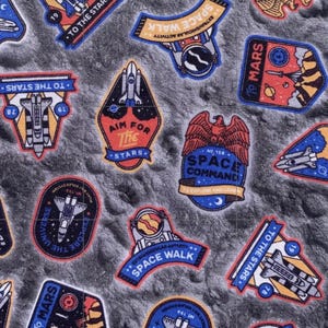 May include: A fabric pattern featuring various space-themed patches. The patches include rockets, planets, and phrases like "Aim for the Stars" and "Space Command." The color palette consists of blues, reds, yellows, and whites against a gray, textured background.