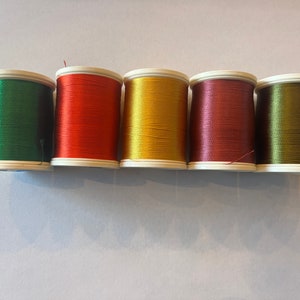 May include: Six spools of thread in different colors: green, orange, yellow, maroon, and olive green. The spools are all the same size and shape, and they are arranged in a row.