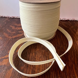 May include: A spool of 100 yards of off-white bias tape. The tape is wound around a cardboard spool with a brown paper label. The tape is lying on a wooden surface.