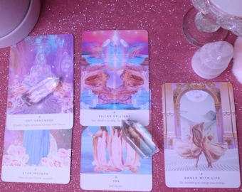May include: Four tarot cards with pastel illustrations and text, including "Get Grounded," "Pillar of Light," "Star Mother," and "Dance with Life." Clear quartz crystals are placed on the cards. The background is a sparkly pink surface.