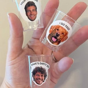 May include: Three clear plastic shot glasses with personalized stickers. The first shot glass has a sticker with a man's face and the text "YOUR TEXT HERE". The second shot glass has a sticker with a dog's face and the text "I DO TOO!". The third shot glass has a sticker with a man's face and the text "DAVE'S DIRTY 30!".