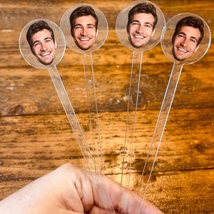 May include: Four clear plastic drink stirrers, each with a circular image of a smiling man's face. The stirrers are held in a hand against a wooden background. The stirrers are transparent and have a long, thin handle.