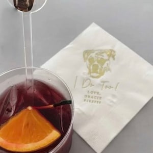 K&ouml;nnte beinhalten: Ein durchsichtiger Plastikbecher gef&uuml;llt mit einem roten Getr&auml;nk und einer Orangenscheibe. Ein durchsichtiger Plastikstab mit einem Foto des Gesichts eines braunen und wei&szlig;en Hundes befindet sich im Becher. Eine wei&szlig;e Serviette mit goldener Aufschrift "I Do Too! Love, Gracie & Brett" befindet sich im Hintergrund.