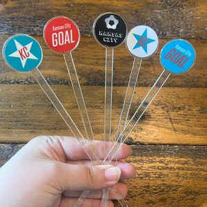 May include: Six clear plastic drink stirrers with round, colorful toppers. The toppers feature a soccer ball, a star, and the text "Kansas City GOAL" and "KC".