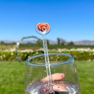 K&ouml;nnte beinhalten: Ein klares Glas gef&uuml;llt mit einem rosa Getr&auml;nk mit Blasen. Ein klarer Plastik-Trinkr&uuml;hrer mit einem runden Aufsatz, der ein gedrucktes Bild eines braunen Hundes zeigt, befindet sich im Glas.