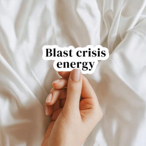 May include: A white sticker with the words "Blast crisis energy" in a black, bold font. The sticker is held by a hand with manicured nails against a white fabric background.
