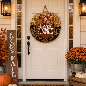 May include: A round door wreath featuring a leopard print border, a central orange circle with the white text "hello PUMPKIN," and a decorative bow. The wreath is hung on a white door alongside a vertical wooden sign reading "hello FALL."