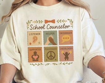 May include: Cream-colored t-shirt with the words "School Counselor" and six illustrated squares. Each square features a different word: Listener, Helper, Encourager, Supporter, Peacemaker, and Cheerleader. The design includes a bow and leaf accents.