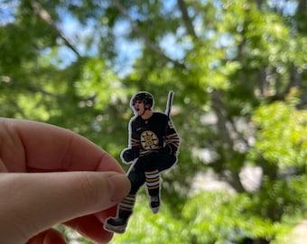 May include: A black and yellow hockey player sticker with the letter "B" on the front. The sticker is held in a hand against a blurred background of green leaves.