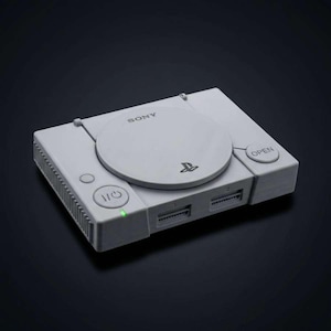 May include: A light gray PlayStation 1 console with the Sony logo and PlayStation symbol. The console has a power button, an open button, and two controller ports. The console is on a black surface.