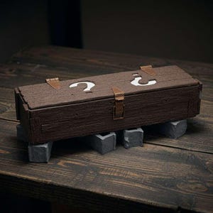 May include: A rectangular dark brown wooden box with a textured wood grain finish. The lid features two white question mark cutouts and is secured with three light brown metal hinges and a central latch. The box rests on four small gray blocks.