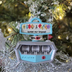 May include: A Christmas tree ornament shaped like a retro arcade game, with the text "PACK A PUNCH" and "increase your firepower!" in colorful letters. The ornament is light blue, gray, and silver, with a red button and a small flag that says "Please Wait".