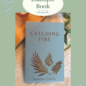 K&ouml;nnte beinhalten: Ein Buch mit dem Titel "Catching Fire" von Suzanne Collins. Das Buch hat einen hellblauen Einband mit goldfarbenen Illustrationen einer Flamme und Bl&auml;ttern. Das Buch wird auf einer strukturierten Oberfl&auml;che mit dem Text "Example Book" oben angezeigt.