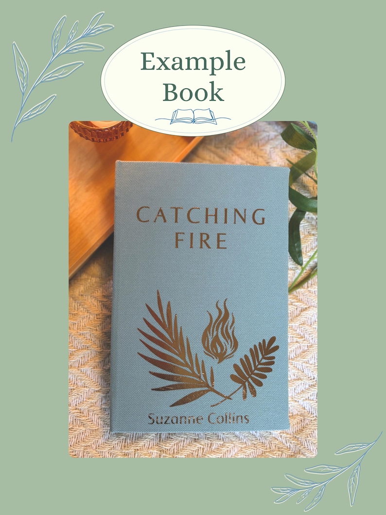 K&ouml;nnte beinhalten: Ein Buch mit dem Titel "Catching Fire" von Suzanne Collins. Das Buch hat einen hellblauen Einband mit goldfarbenen Illustrationen einer Flamme und Bl&auml;ttern. Das Buch wird auf einer strukturierten Oberfl&auml;che mit dem Text "Example Book" oben angezeigt.
