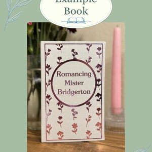 K&ouml;nnte beinhalten: Ein Buch mit dem Titel "Romancing Mister Bridgerton" mit einem bordeauxfarbenen Blumendesign. Das Buch liegt auf einer Holzoberfl&auml;che, mit einer rosa Kerze und einer Vase mit Blumen im Hintergrund. Der obere Teil des Bildes tr&auml;gt den Text "Example Book".