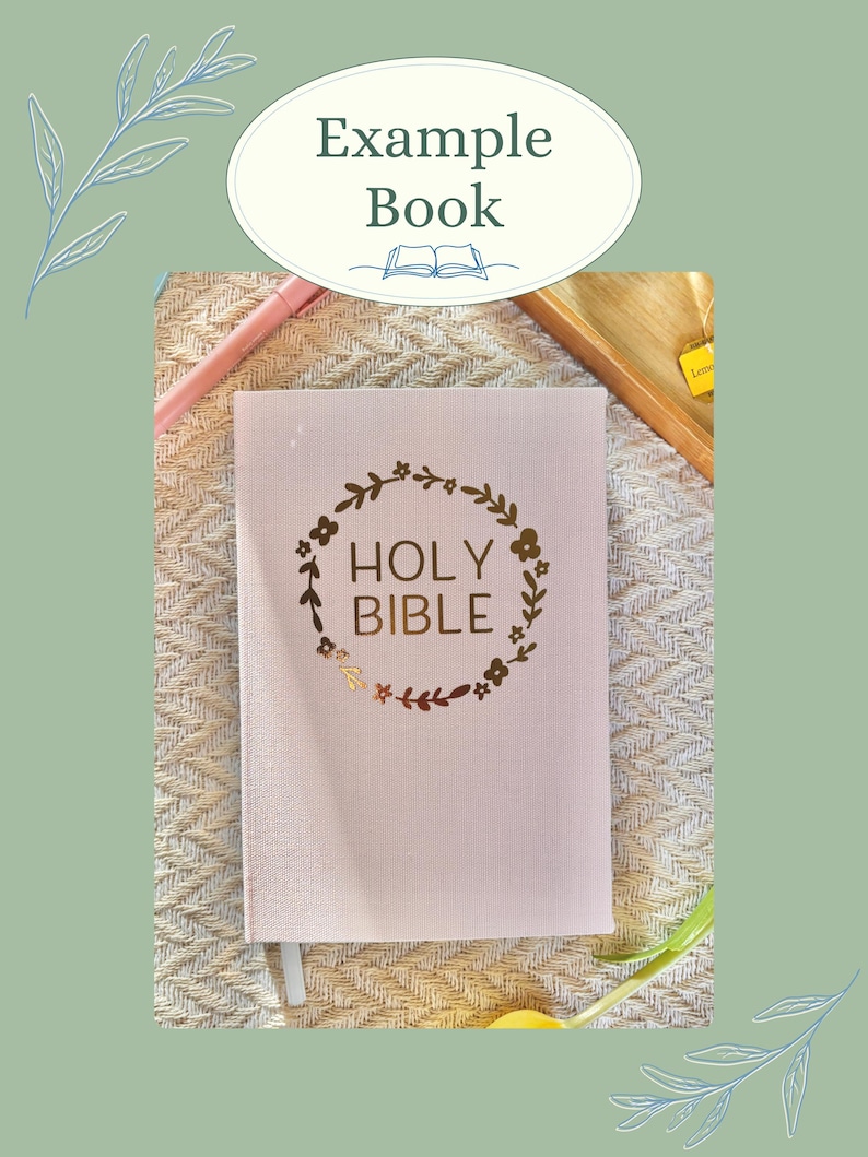 K&ouml;nnte beinhalten: Ein hellrosa Heilige Bibel mit goldfarbenen Buchstaben und einem floralen Kranzdesign. Das Buch hat ein Leseband und liegt auf einer strukturierten Oberfl&auml;che. Das Bild enth&auml;lt auch den Text "Example Book".