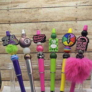May include: A collection of decorative pens with various embellishments. The pens feature colorful designs, including text that reads "I'm not like other girls", "What kind of Fuckery is this?", and "Little Miss Fucking Sunshine". The pens are displayed on a clear stand.