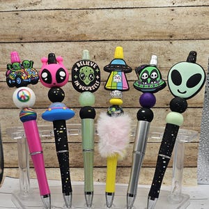 May include: A collection of decorative pens with unique designs. Each pen features a different topper, including aliens, UFOs, and the words "Believe in Yourself." The pens are displayed on a clear stand, showcasing their colorful and whimsical details.