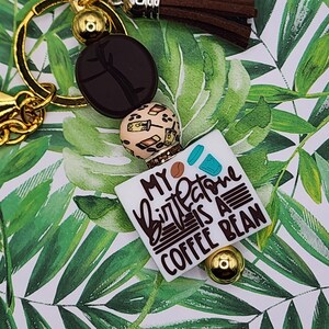 May include: Gold keychain with a brown tassel, a brown coffee bean charm, and a white charm with the text "My  Buttone is a Coffee Bean".