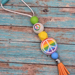 Peut inclure: Un collier de perles en silicone coloré avec un pendentif en forme de signe de paix. Les perles sont vertes, blanches, jaunes et bleues. Le pendentif en forme de signe de paix a un motif arc-en-ciel. Le collier a un pompon à l'extrémité.
