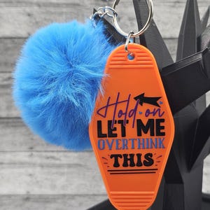 May include: An orange motel-style keychain with the text "Hold on, Let me overthink this" in blue and black. It is attached to a silver keyring and a fluffy blue pom-pom.