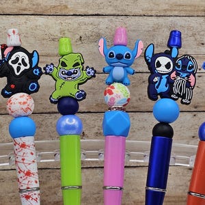 Puede incluir: Una colección de bolígrafos coloridos con adornos temáticos de personajes. Los bolígrafos presentan diseños como Stitch, una cara de fantasma y un monstruo verde. Los barriles de los bolígrafos son de varios colores y algunos tienen cuentas decorativas. El bolígrafo azul tiene el texto "OHANA MEANS FAMILY".
