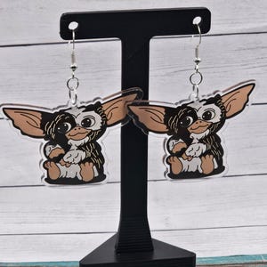 May include: A pair of dangle earrings featuring a cartoon character with black fur, white eyes, and brown ears. The earrings are made of clear acrylic and have silver hooks.