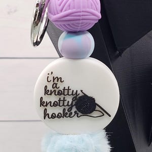 May include: A keychain with a purple yarn ball, a light blue and purple bead, and a white disc with the text "I'm a knotty knotty hooker" and a black yarn and crochet hook design. A light blue pom-pom is at the bottom.