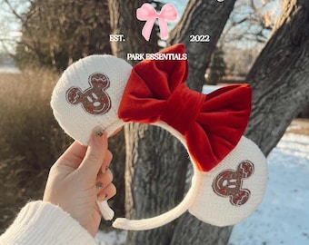 May include: A white knit headband featuring two circular ears with embroidered brown gingerbread mouse-shaped cookies. A large, vibrant red velvet bow is centered between the ears. The text sarah belle magic, EST. 2022, and PARK ESSENTIALS is visible.