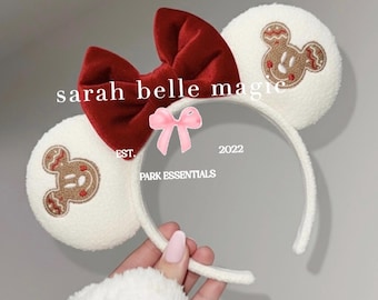 May include: White headband with two round, textured ears featuring embroidered gingerbread Mickey Mouse designs. A large, red velvet bow sits between the ears. The text "sarah belle magic" and "PARK ESSENTIALS" are visible.