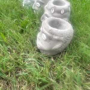 May include: Three gray concrete planters shaped like human faces with white seashells for eyes and a green beaded headband on the top planter.