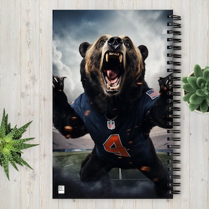May include: Spiral-bound notebook featuring a digitally enhanced image of a roaring bear wearing a navy blue football jersey with the number 4. The bear is set against a stadium backdrop with a cloudy sky.