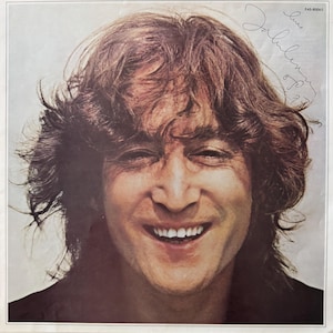 Pode incluir: Uma fotografia preto e branco de John Lennon sorrindo com o cabelo cobrindo a testa. A foto é assinada "John Lennon" com a data "6/71" e o número "EAS-80045".