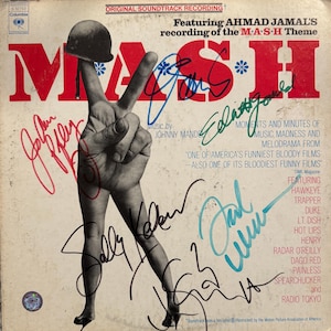 Może przedstawiać: Vintage okładka albumu ze ścieżką dźwiękową MASH. Grafika albumu przedstawia dłoń robiącą znak pokoju z hełmem na palcu wskazującym. Słowo "MASH" jest dużymi czerwonymi literami. Okładka zawiera podpisy i tekst.