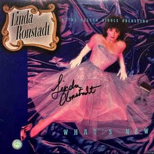 May include: Album cover for Linda Ronstadt's "What's New" featuring a glamorous woman in a pink strapless gown. The album title and artist's name are prominently displayed, with a signature and the text "WHAT'S NEW". The background is a mix of blue and purple.