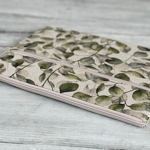 May include: A beige zippered pouch with a double zipper closure. The pouch is decorated with a green and white floral pattern. The pouch is made of fabric and has a soft, textured feel.