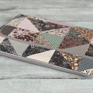 Può includere: Una pochette grigia con cerniera con un design a patchwork che presenta vari motivi floreali in tonalità di rosa, marrone, verde e grigio.