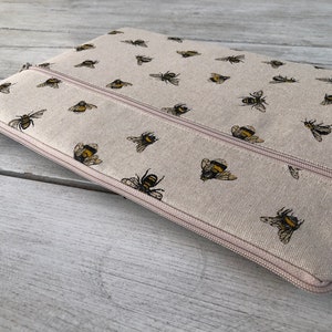 Puede incluir: Una bolsa beige con cremallera que tiene una doble cremallera. La bolsa está hecha de una tela similar al lino y presenta un patrón repetitivo de abejas negras y amarillas.