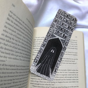 May include: Black and white bookmark featuring a ghost in front of a building with many windows. The building is filled with bookshelves. The bookmark has a dotted texture.