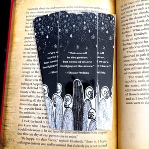 Può includere: Tre segnalibri in bianco e nero con una citazione di Oscar Wilde, un design a cielo stellato e illustrazioni di figure con volti a forma di teschio. I segnalibri sono all'interno di un libro aperto con pagine invecchiate.