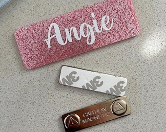 May include: A pink glitter name tag with the name "Angie" in white script. Includes a 3M adhesive strip and a magnetic name tag backing with the text "CAUTION MAGNETIC".